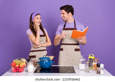 Photo Of Funny Positive Husband Wife Wear Grey Aprons Reading Pointing Cook Book Isolated Violet Color Background