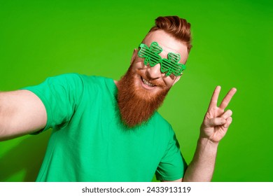 Photo of funny positive man with ginger beard in clover glasses showing v-sign symbol make selfie isolated on green color background