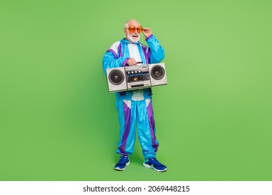 Photo of funny pretty senior man wear sportive suit dark eyewear listening boombox smiling isolated green color background