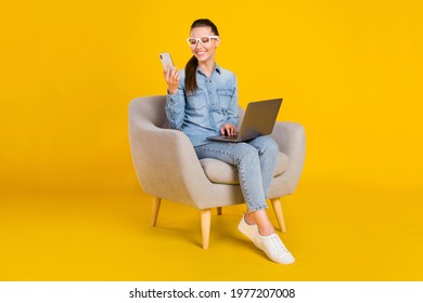 Photo of funny pretty young woman wear denim shirt glasses smiling sitting chair typing modern gadgets isolated yellow color background