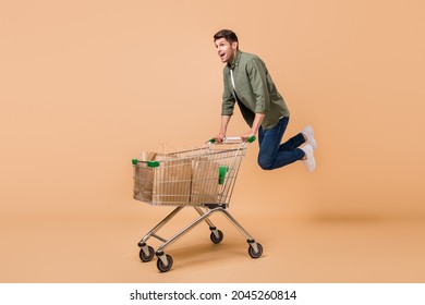 Photo of funny sweet young guy dressed green shirt smiling doing purchases riding shopping trolley isolated beige color background