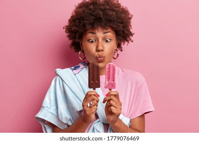 Photo of funny teenage girl keeps lips rounded and looks at appetizing ice cream, enjoys summer time, likes cold dessert, dressed in stylish outfit, chooses between chocolate and strawberry flavor