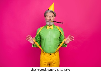 Photo of funny white haired grandpa hold yellow suspenders blow noisemaker funky crazy birthday party surprise wear paper cap green shirt pants isolated bright pink color background
