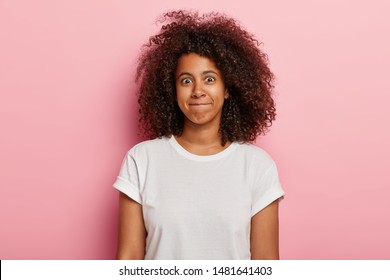Photo of funny woman has curly thick hair, presses lips together, has happy face, wears white t shirt, isolated over pink background. Good looking young African American girl expresses happiness.