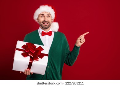 Photo of gentleman wear green cardigan bowtie santa hat hold box with bun finger point new shopping exclusive brand isolated on red color background