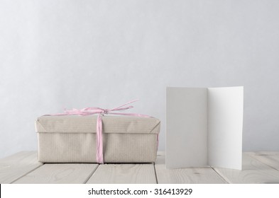 Photo Of A Gift Box Wrapped In Brown Paper, Tied With Pale Icy Pink Raffia On An Old Planked Wooden Table. A Blank Open Greeting Card Faces Front  Retro Style With Pale Pastel Hues.