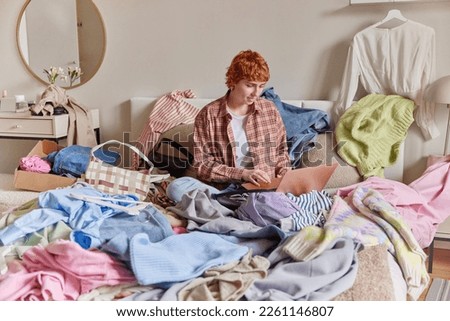 Photo of ginger woman makes revision of clothing at home sits on messy bed uses laptop computer sells different outfits to wear dressed in checked shirt poses in bedroom surrounded by lots of clothes