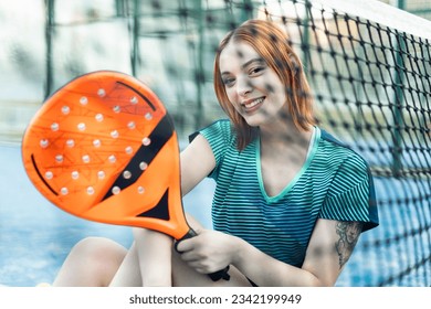 Photo of a girl behind a net of an outdoor paddle tennis court who is sitting looking at the camera happily with a racket. Concept of women playing paddle. Paddle for women.
