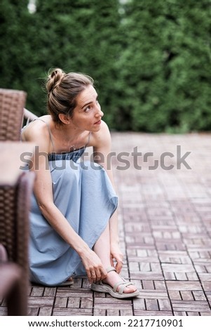 Photo of a girl sitting in a wicker chair on a green terrace, she is dressed in a long loose blue dress. Emotions. Outdoor recreation. Reflections on oneself. Lady in the modern city.