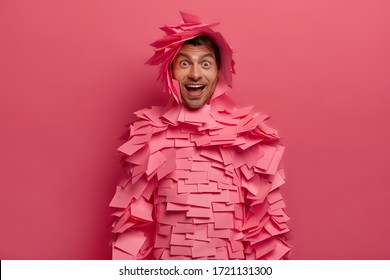 Photo of glad positive woman looks with wonder and happiness at camera, has playful mood, giggles over funny joke, makes paper outfit of stickers, isolated on pink background, has hilarious talk