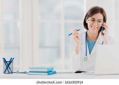 Photo Of Glad Woman Therapist Or Physician Has Phone Conversation With Patient, Gives Advice How To Cure Disease, Wears White Gown, Reads Information On Laptop Computer, Works In Private Clinic