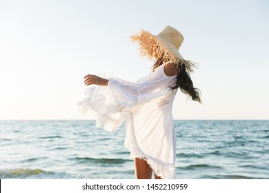 Photo Of Glamorous Brunette Woman In Beach Dress And Straw Hat Walking By Seaside In Summer Morning