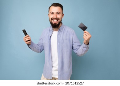 Photo of good looking attractive smiling brunet unshaven young man wearing casual blue shirt and white t-shirt isolated over blue background wall holding credit card and mobile phone