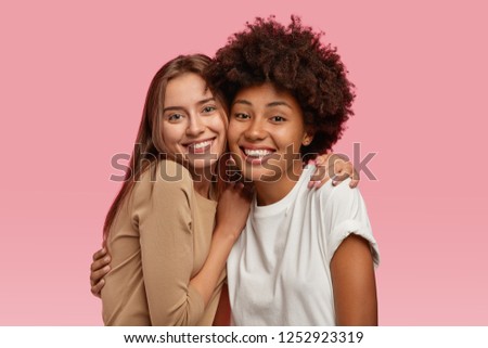 Photo of good looking two young models stand closely, embrace and enjoy togetherness, dressed in casual clothes, model over pink background. People, interracial friendship, happiness concept