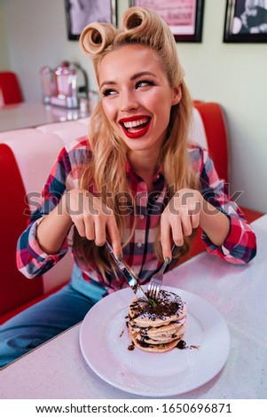 Photo of gorgeous cheerful woman with beautiful hairstyle eating pancakes and laughing in retro american cafe