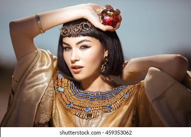 Photo Of Gorgeous Woman With Cleopatra Makeup, Closeup Portrait Of Beautiful Female With Stylish Haircut Agaist, Young Lady Wearing Fashionable Golden Necklace And Holding Magik Ball Outdoor, Beauty