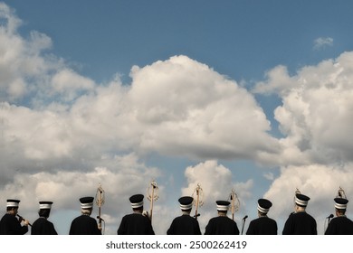 In the photo, a group of musicians with their backs turned are looking towards the sky with their instruments. White clouds and the blue sky contrast with the musicians' traditional attire. - Powered by Shutterstock