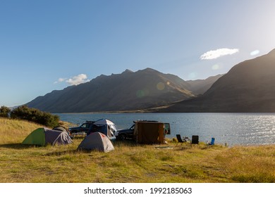 A photo of a group of tents put up by the lake. Two unrecognizable cars standing sideways behind the tents. Mavora Lake and Livingstone Mountain range in the background. Sunny, summer day before sunse - Shutterstock ID 1992185063