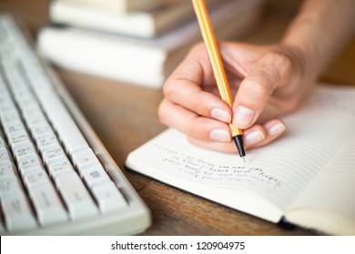 Photo of hands writes a pen in a notebook, computer keyboard and a stack of books in background