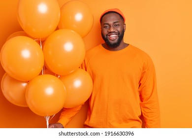 Photo of handsome bearded dark skinned man smiles joyfully dressed in casual clothes holds bunch of inflated balloons celebrates specical occasion isolated over orange background. Festive concept