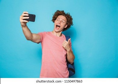 Photo Of Handsome College Guy With Curly Hair Wearing Casual Clothing And Backpack Holding Smartphone And Taking Selfie Isolated Over Blue Background