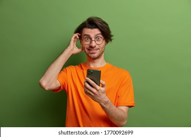 Photo of handsome hipster guy with trendy hairstyle, scratches head and thinks about something, uses modern mobile phone, checks newsfeed, dressed in orange t shirt, isolated on green background - Powered by Shutterstock