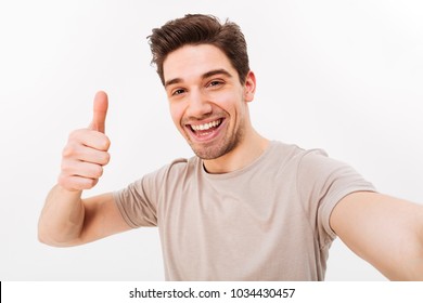 Photo of handsome man in casual t-shirt and bristle on face smiling on camera with thumb up while taking selfie isolated over white background