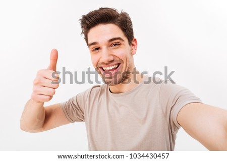 Photo of handsome man in casual t-shirt and bristle on face smiling on camera with thumb up while taking selfie isolated over white background