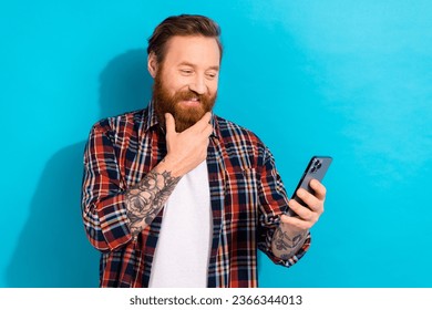 Photo of handsome man with stylish beard dressed checkered shirt look at smartphone hand on chin isolated on blue color background - Shutterstock ID 2366344013