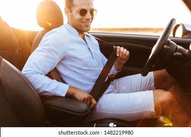 Photo Of Handsome Man In Sunglasses Wearing Seat Belt While Driving Convertible Car By Seaside At Sunrise