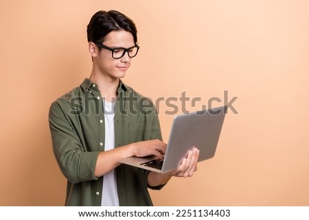 Photo of handsome positive cheerful guy with brunet hair dressed khaki shirt typing chatting on laptop isolated on beige color background