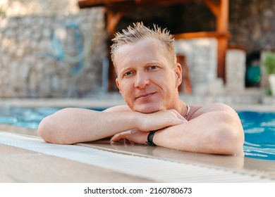 Photo Of Handsome Smiling Man In Swimming Pool In Summer Scenery. Attractive, Cheerful, Stunning, Perfect, Harsh, Virile Man In Yellow Shorts, Having Cocktail With Small Umbrella In Hand And Holding