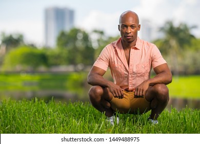 Photo Of A Handsome Young African American Man Squatting On Grass In The Park. Deadpan Expression Staring Deep Into The Camera