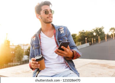 Photo of handsome young guy walking outdoors using mobile phone drinking coffee listening music. - Powered by Shutterstock