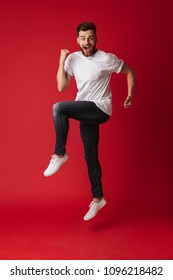 Photo of handsome young man jumping isolated over red wall background showing winner gesture.