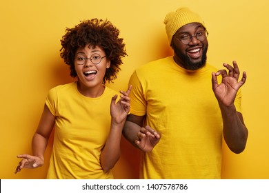 Photo Of Happy African Couple Dance Together Against Yellow Background, Move Body Actively, Show Okay Gesture, Wear Casual Yellow T Shirt, Have Fun During Party. Monochrome. Feeling Rhytm Of Music