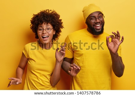 Photo of happy African couple dance together against yellow background, move body actively, show okay gesture, wear casual yellow t shirt, have fun during party. Monochrome. Feeling rhytm of music