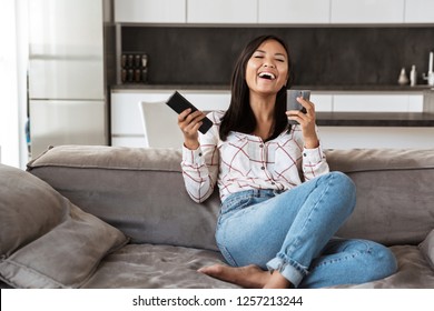 Photo of happy asian woman 20s holding remote control and cup while sitting at sofa in cozy apartment