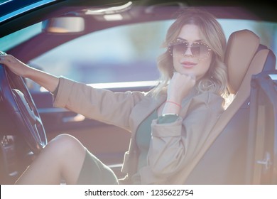 Photo of happy blonde with glasses and long dress sitting in car with open door