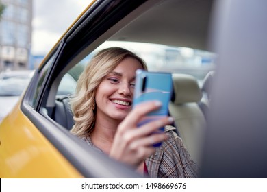 Photo of happy blonde with phone in her hand sitting in back seat in yellow taxi