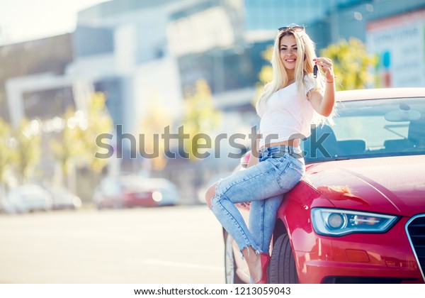 Photo of happy blonde woman with keys standing near
red car on summer day.