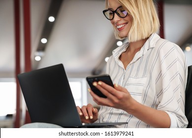 Photo of happy blonde woman wearing eyeglasses using cellphone and laptop while sitting in conference hall
