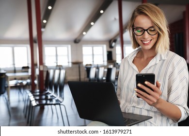 Photo of happy blonde woman wearing eyeglasses using cellphone and laptop while sitting in conference hall