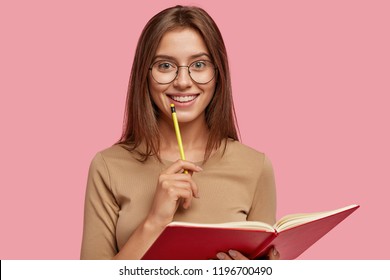 Photo of happy brunette woman with positive smile, carries textbook, holds pencil for writing, makes notes while listens some information, poses over pink background. Its time for creating new poem! - Powered by Shutterstock