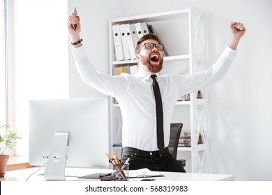 Photo Of Happy Businessman Standing In Office Near Computer Make Winner Gesture.