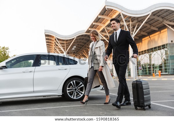 Photo of happy\
colleagues man and woman in formal wear walking with suitcase on\
parking near airport\
outdoors