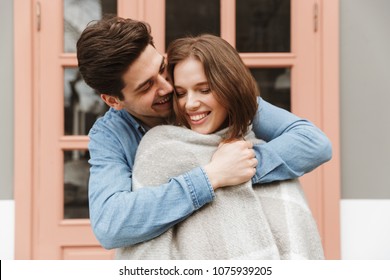 Photo Of Happy Couple In Love Standing Outside And Man Wrapping His Woman In Warm Blanket