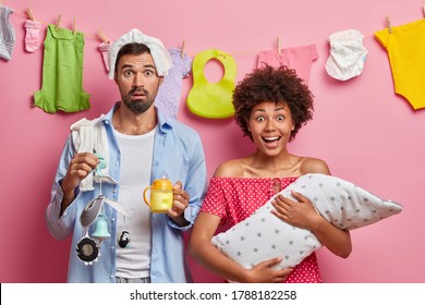 Photo Of Happy Dark Skinned Mother Holds Small Baby Wrapped In Blanket, Shocked Father With Nappy On Head, Feeding Bottle And Mobile, Busy Nursing Infant, Washed Newborn Clothes In Background