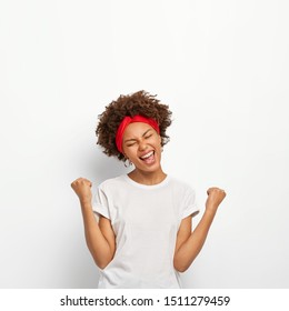 Photo of happy delighted Afro girl clenches fists, feels triumph, rejoices victory, keeps eyes shut, smiles broadly, wears white t shirt, stands indoor. Yes, I did it successfully. People, high spirit