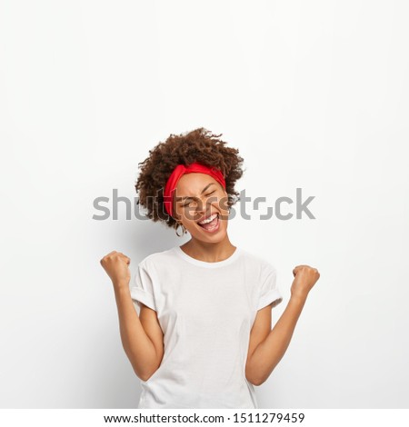Photo of happy delighted Afro girl clenches fists, feels triumph, rejoices victory, keeps eyes shut, smiles broadly, wears white t shirt, stands indoor. Yes, I did it successfully. People, high spirit
